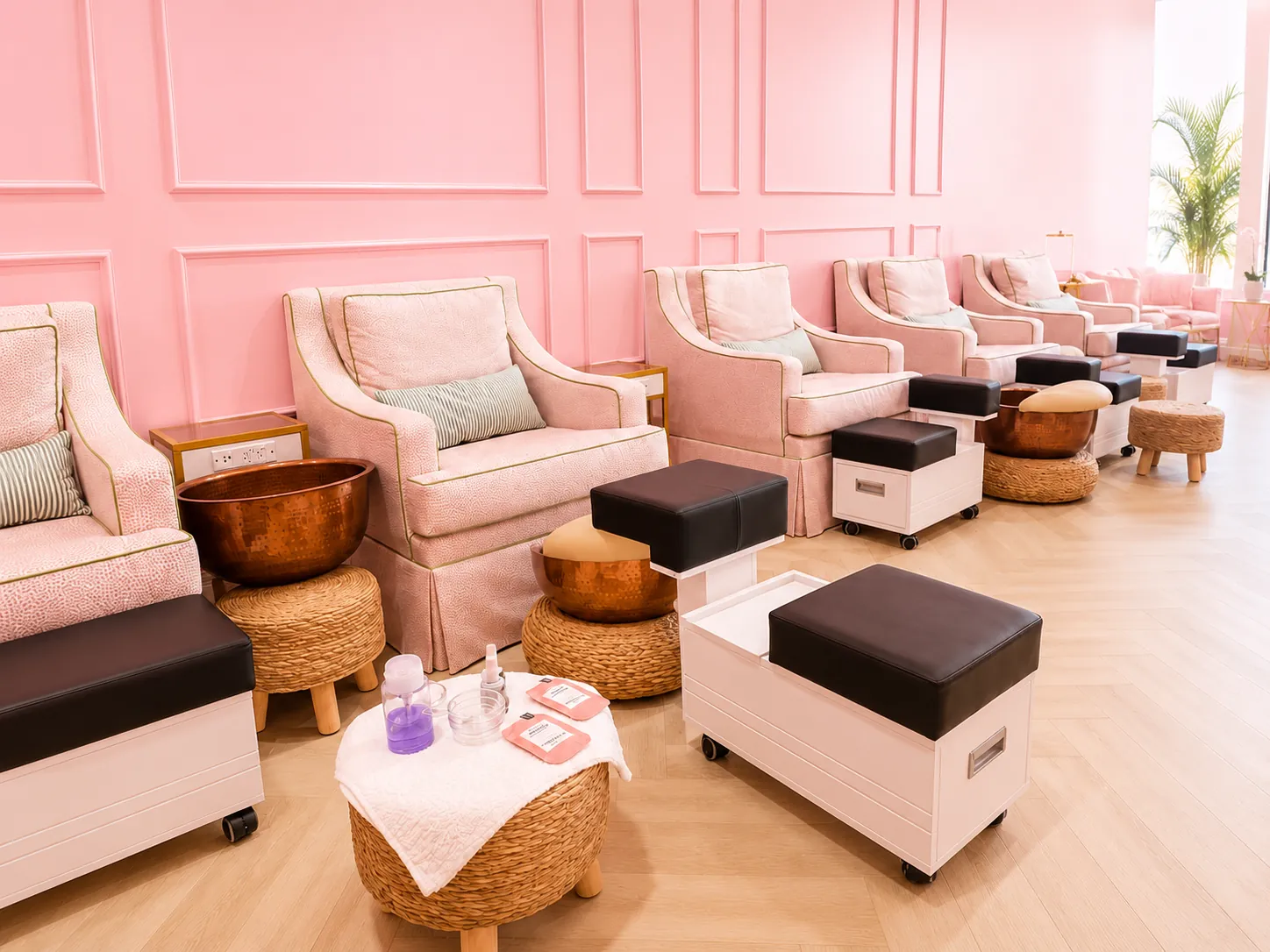 Row of pink pedicure chairs with hammered copper bowls and woven stools against a pink paneled wall