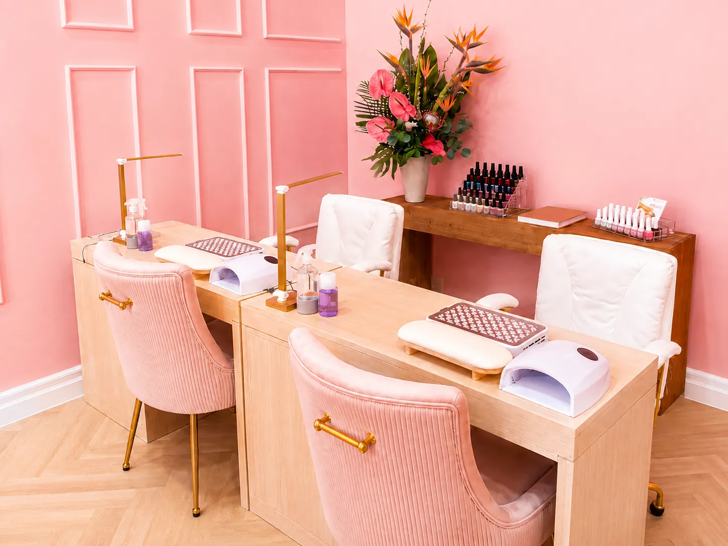 Two Palm Nail Lounge manicure stations with pink velvet chairs, gold lamps, and a tropical floral arrangement