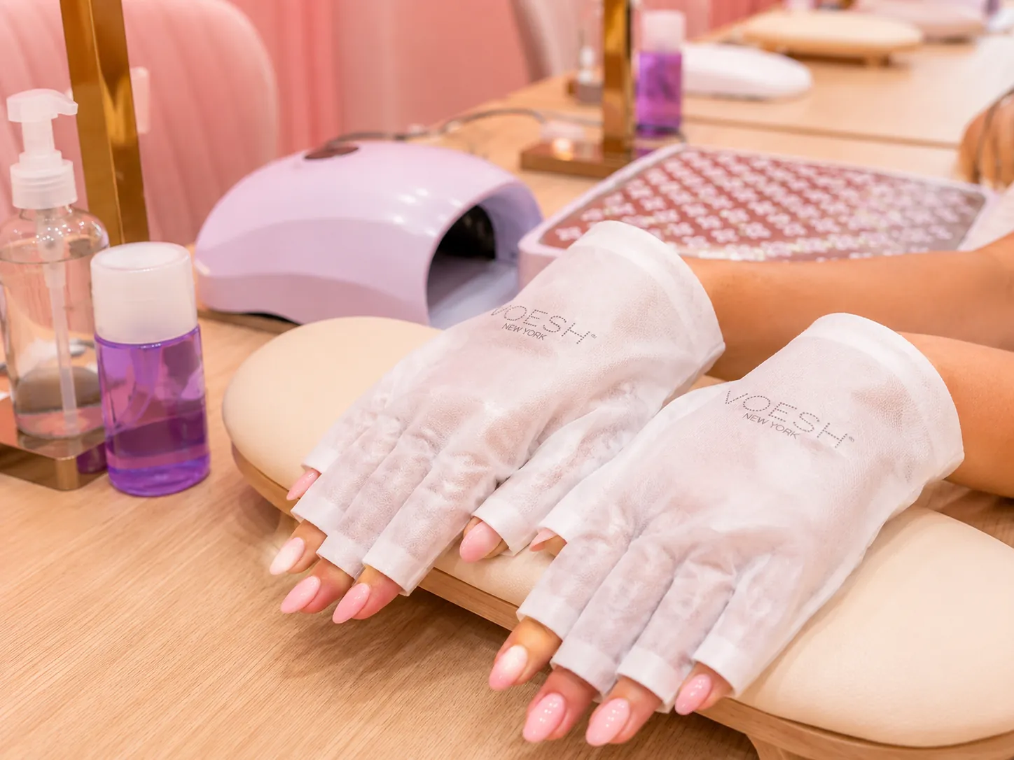 Hands with pink almond nails resting in UV-protective gloves beside a lilac curing lamp