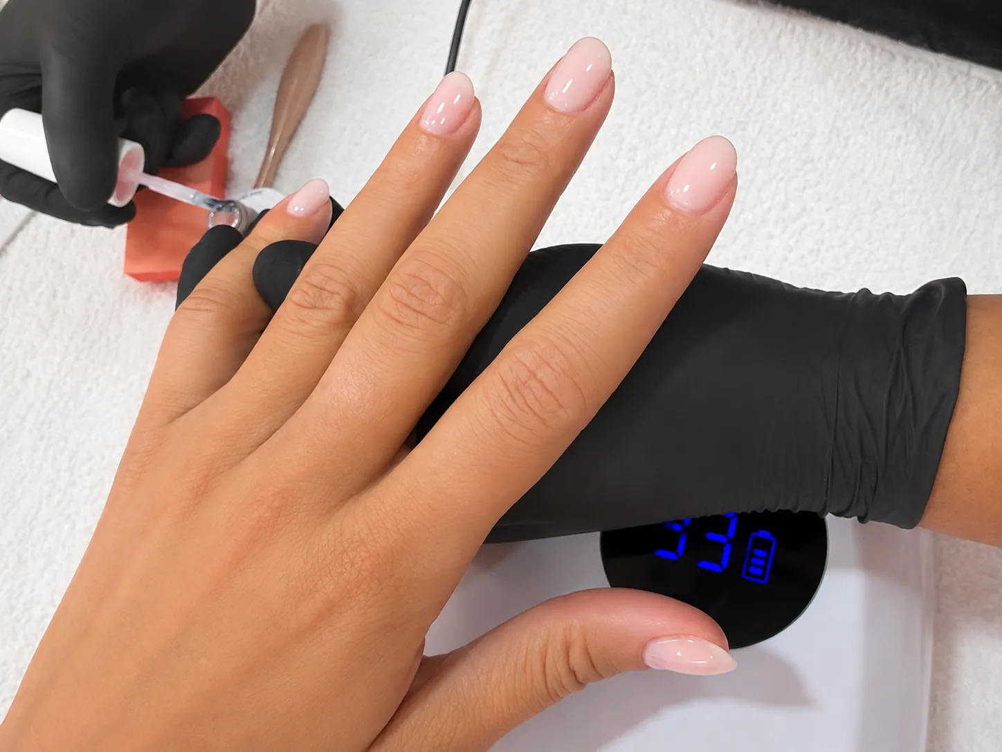 A technician in black gloves applying finishing product to a soft pink almond Russian manicure under a curing lamp